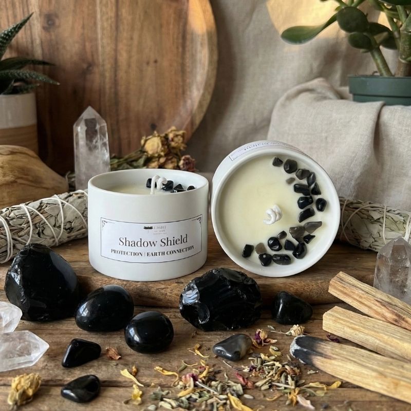 Candle labeled 'Shadow Shield' surrounded by black stones, herbs, and wooden sticks on a wooden surface.