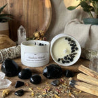 Candle labeled 'Shadow Shield' surrounded by black stones, herbs, and wooden sticks on a wooden surface.